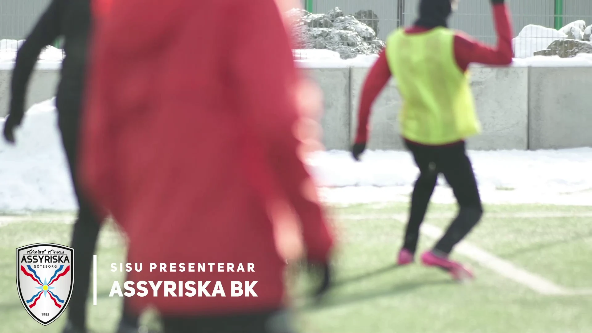 Fotbollsträning på en snöig konstgräsplan, med texten ”SISU presenterar Assyriska BK” och klubbens märke.
