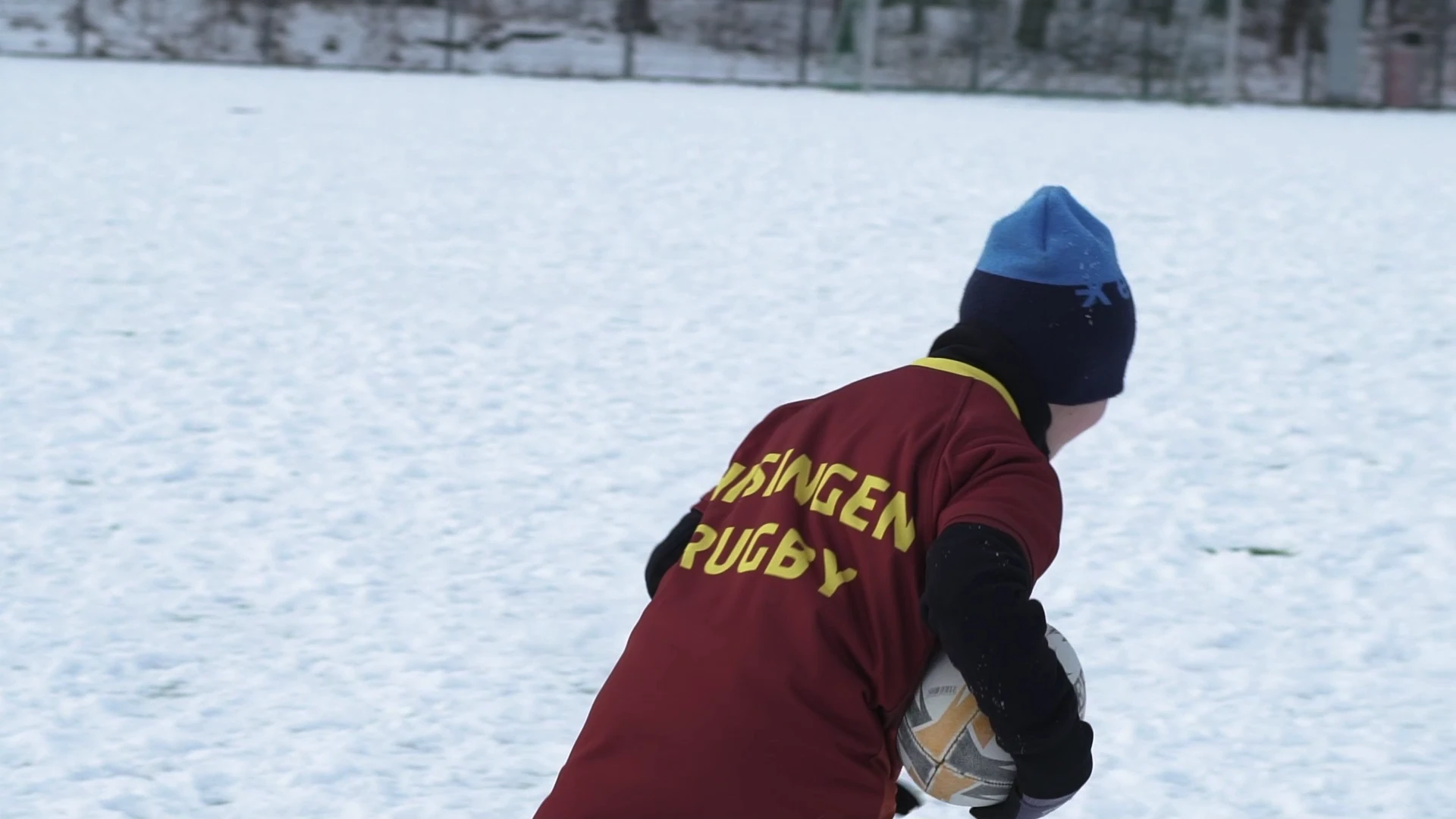 En rugbyspelare i Hisingens rödbruna tröja håller bollen på en snötäckt plan, sedd bakifrån.