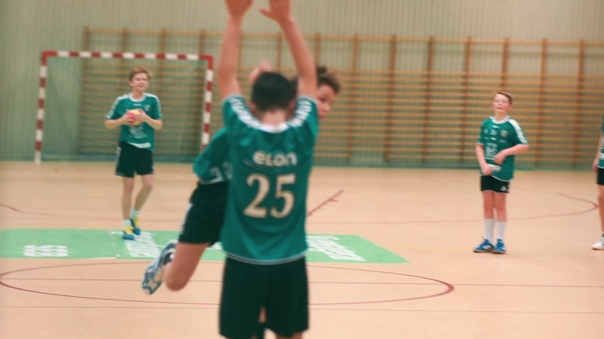 En handbollsspelare med nummer 25 på den gröna tröjan hoppar med armarna uppsträckta, med lagkamrater och ett mål i bakgrunden.