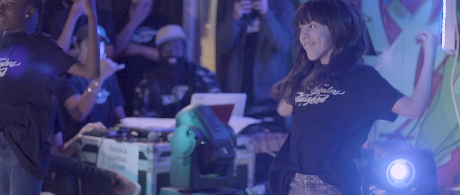 A young woman dances by a stage with DJ decks, with a spotlight shining in the foreground.