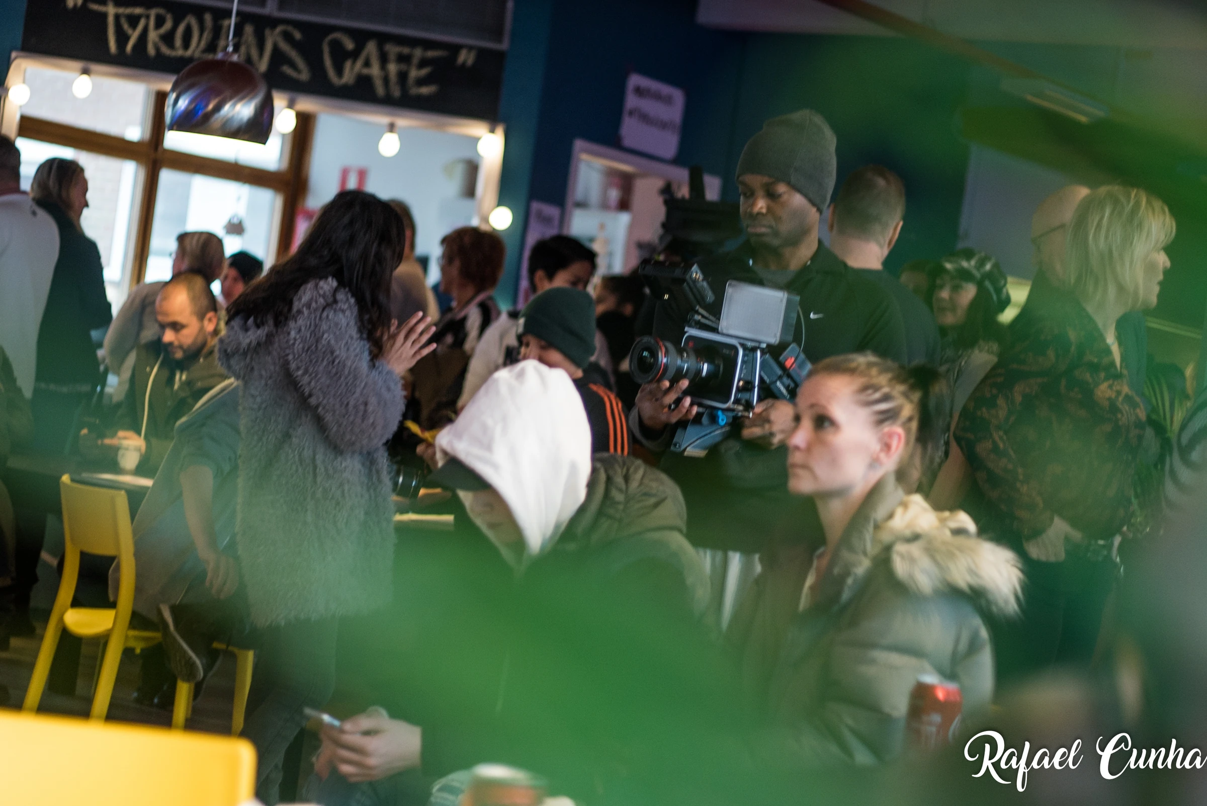 A camera operator with a film camera stands among the visitors in a full café, with the sign “Tyrolens Café” on the wall.