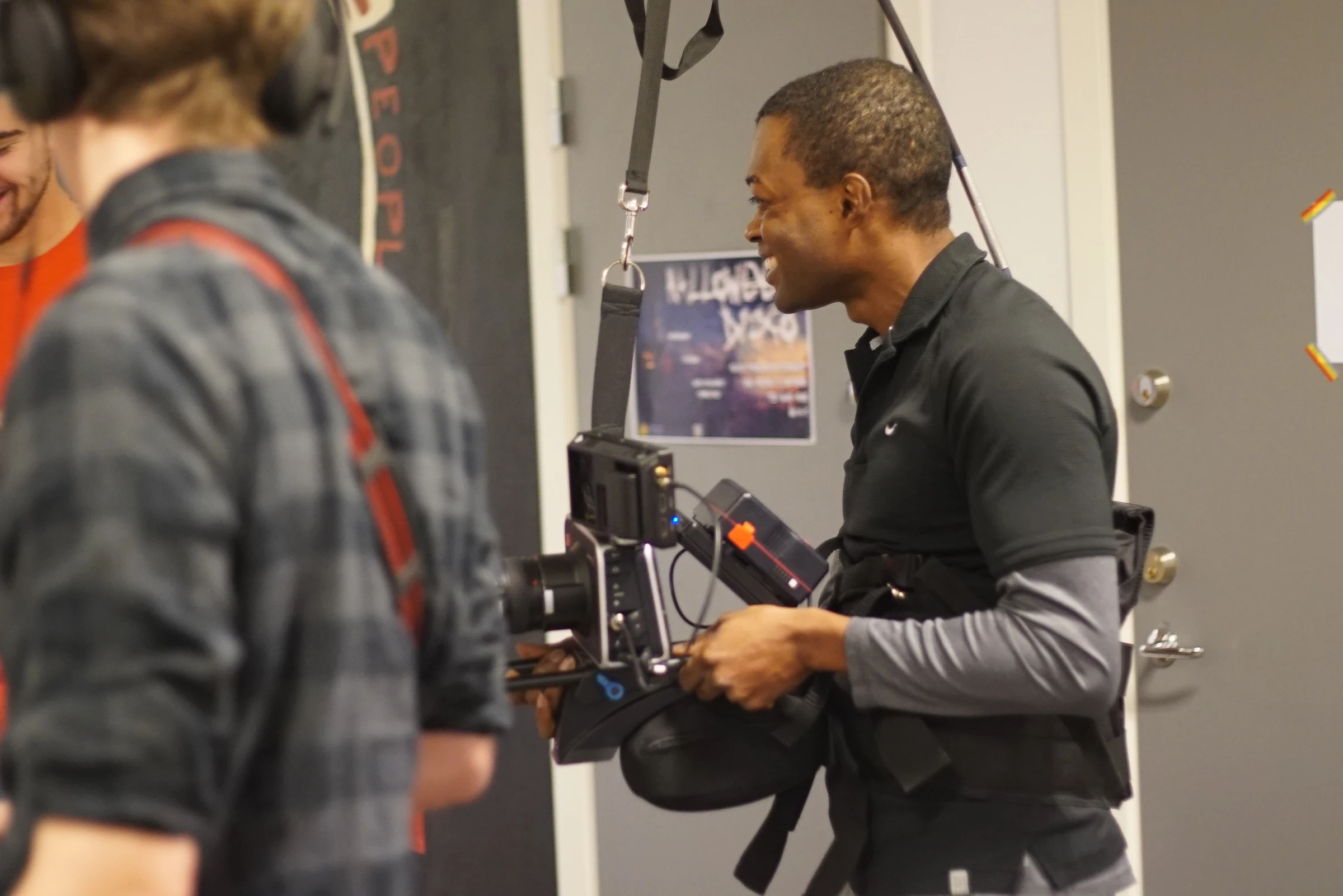 A camera operator with the camera on a support harness talks to a colleague in a corridor, with a sound technician in headphones in the foreground.