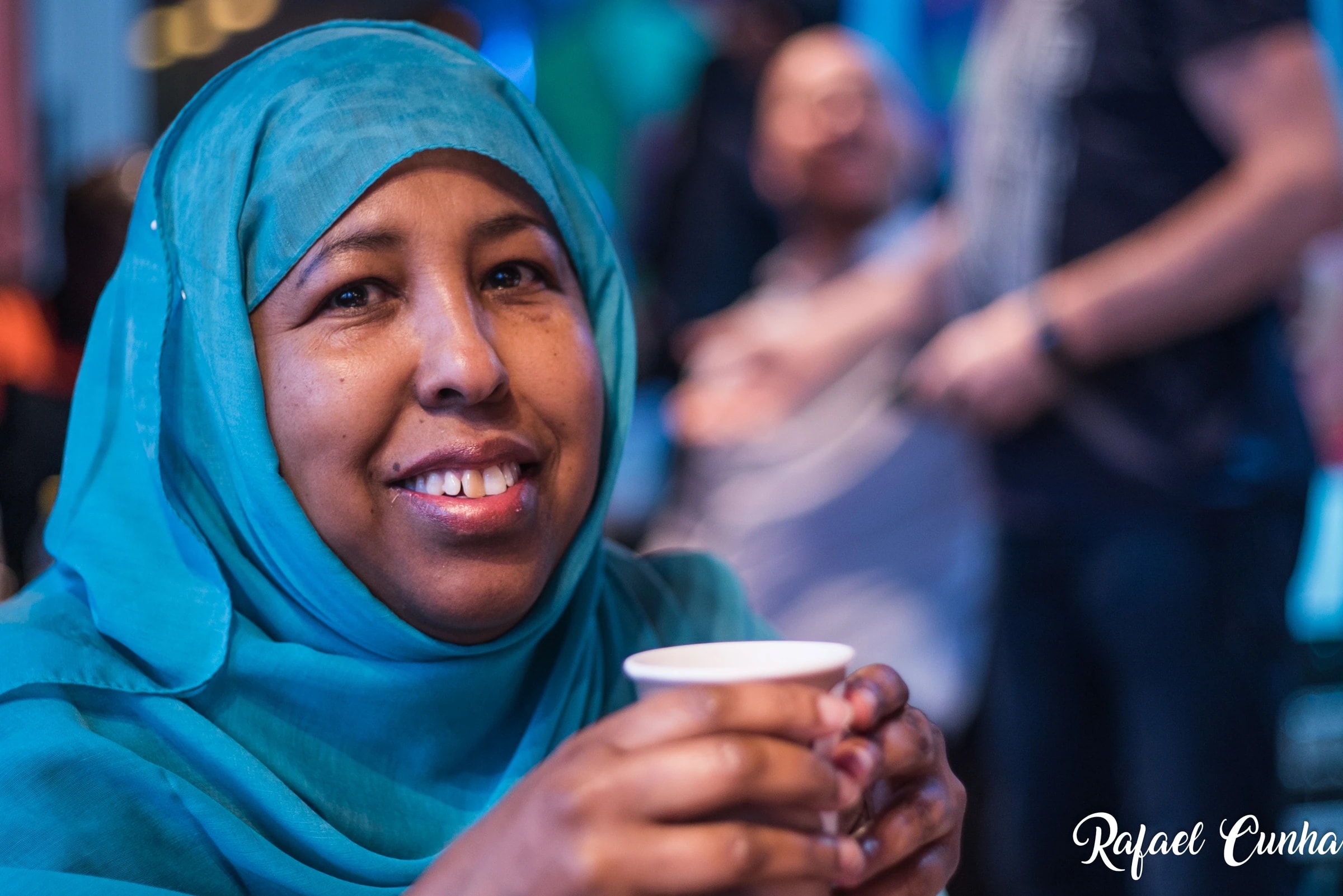 A woman in a turquoise headscarf holds a coffee cup and smiles.