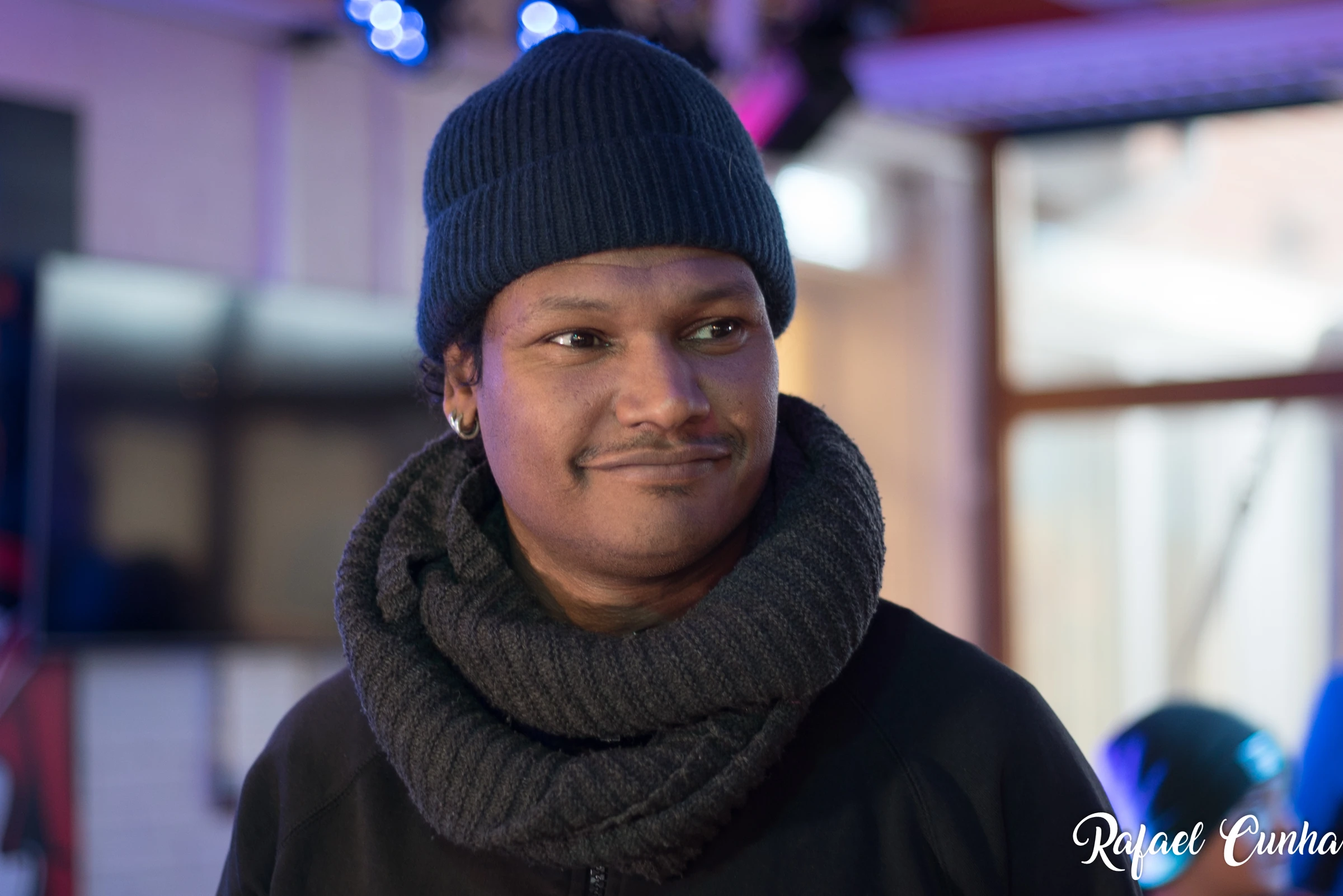A man in a dark beanie and a thick scarf smiles and looks to the side.