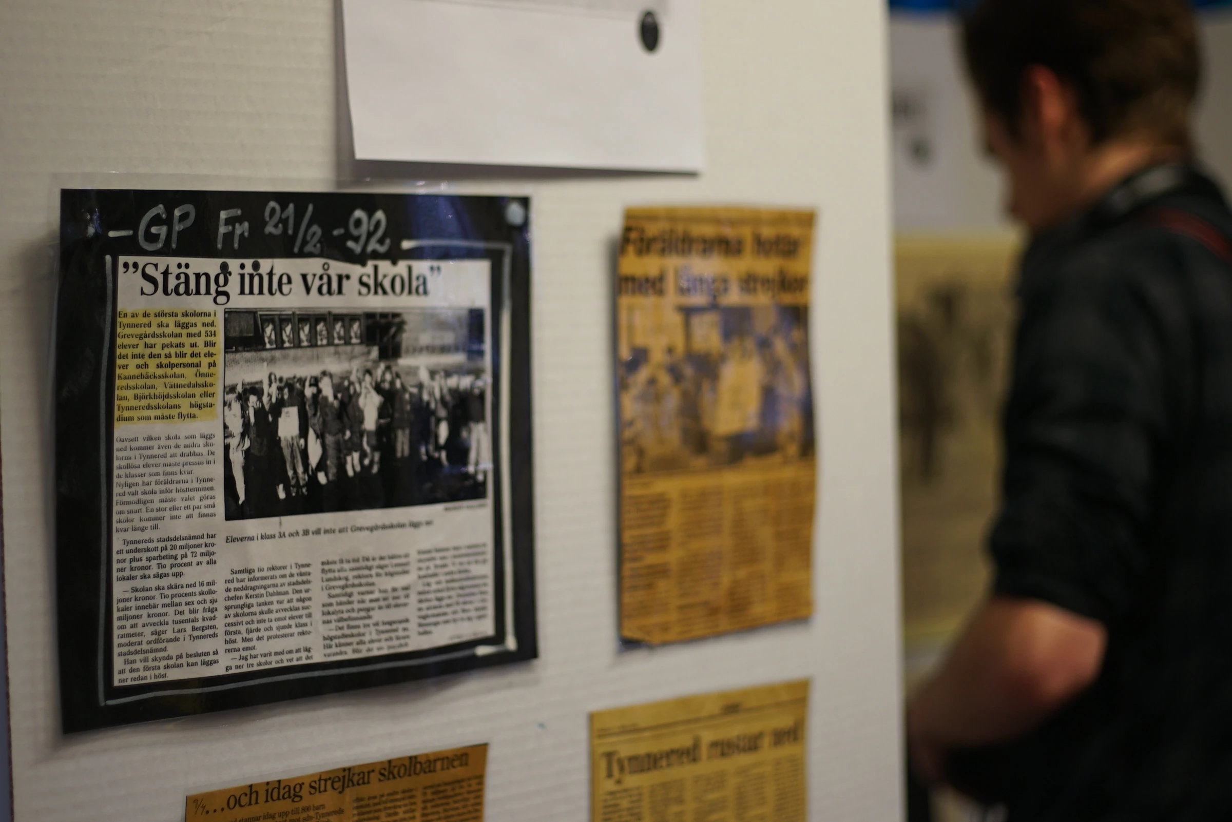 A framed newspaper clipping with the headline “Stäng inte vår skola” hangs on a wall next to other old clippings.