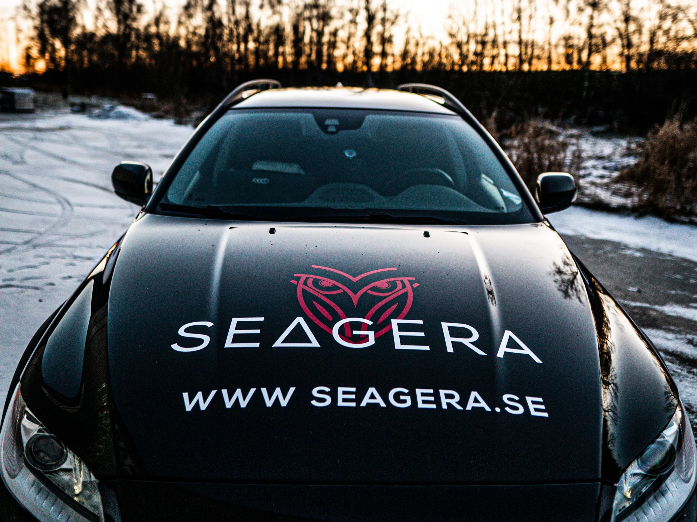 A black car in snow at sunset, with a red owl, the word SEAGERA and www.seagera.se in white on the bonnet.