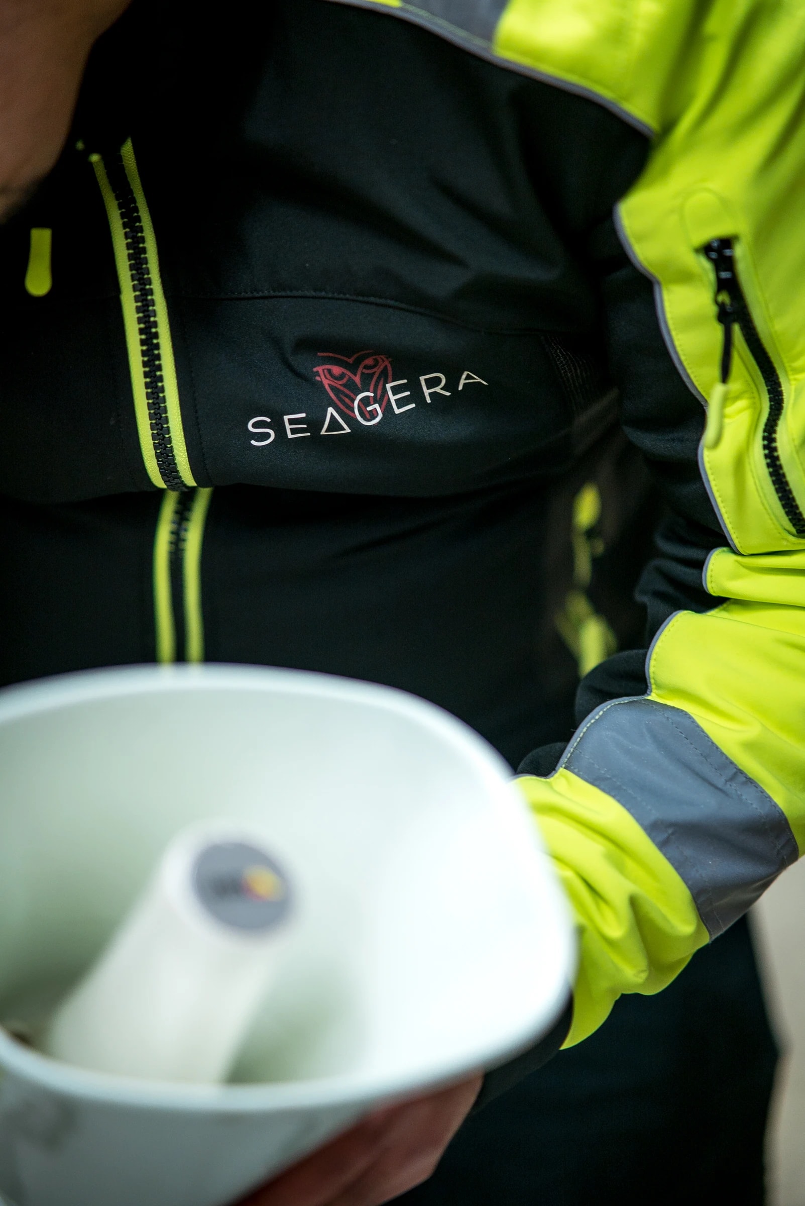 Close-up of a black and yellow work jacket with the owl and SEAGERA on the chest, with a white horn speaker in the foreground.